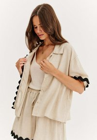 Béžová lněná košile s krátkým rukávem a černým zakulaceným lemem, zapnutá, nošená přes světlý top na ramínka, spárovaná s odpovídajícími světlými šortkami.