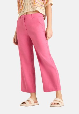 Femme portant un pantalon large rose vif, un haut en crochet crème et des sandales tressées crème, debout sur un fond blanc.