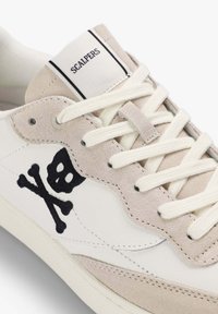 Zapatilla blanca y beige con cordones blancos, etiqueta "Scalpers" en la lengüeta y diseño de calavera y huesos cruzados en negro en el lateral.
