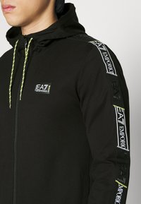 Uomo che indossa una giacca a zip nera con cappuccio, con cordini gialli e logo EA7 Emporio Armani sul petto e sulle maniche.