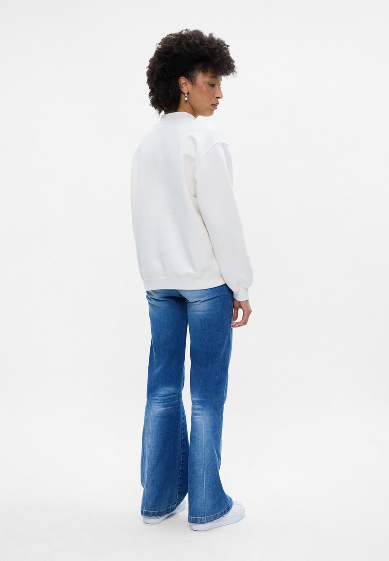 Sudadera blanca con puños y dobladillo acanalados, combinada con jeans acampanados de color azul claro. Accesorios de pendientes de plata, la modelo se encuentra frente a un fondo neutro.