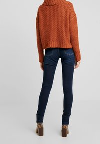 Stickad orange tröja med hög krage, matchad med mörkblå smalbyxor och bruna högklackade stövlar med ormskinnsmönster.