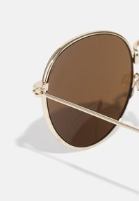 Lunettes de soleil à monture dorée avec de grandes verres bruns. La monture présente un design épuré et un nez bien marqué, avec des branches fines.