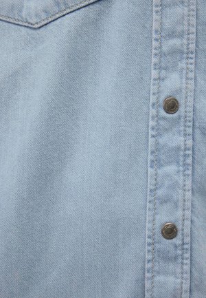 Tessuto di denim azzurro chiaro con grana verticale, caratterizzato da una fila di bottoni metallici lungo il lato destro e dettagli di cucitura visibili.