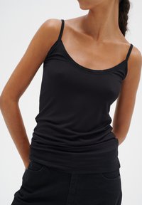 Camisola nera attillata con spalline sottili, caratterizzata da una texture morbida e liscia e un collo rotondo. Indossata con pantaloni scuri.