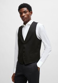 Svart skreddersydd vest med V-hals design, med fire svarte knapper, to framlommer, og glatt stoff, brukt over en hvit skjorte.