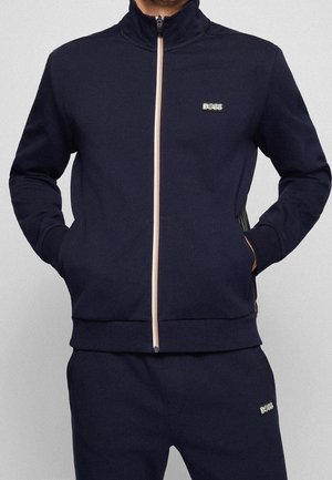 Uomo in giacca a zip e pantaloni coordinati di tuta blu navy con il logo BOSS sul petto e sulla coscia, mani nelle tasche della giacca.