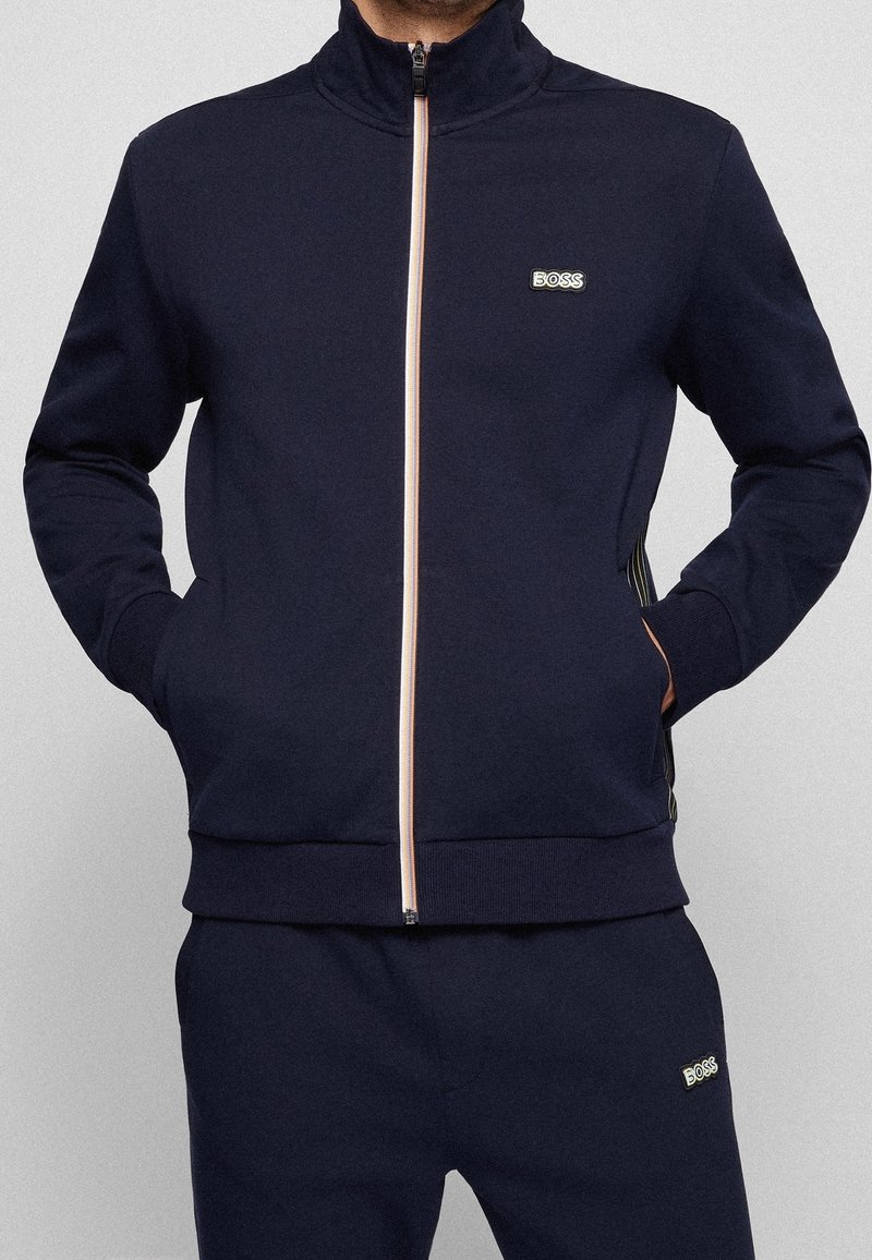 Man som bär en marinblå zip-up träningsjacka och matchande byxor med BOSS-logotyp på bröstet och låret, med händerna i jackfickorna.