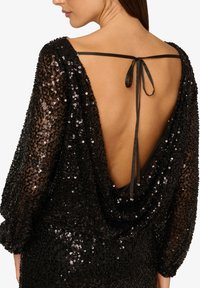 Czarna sukienka z cekinami, z głębokim dekoltem na plecach, luźnymi rękawami i wiązaną wstążką z tyłu. Teksturowany i błyszczący materiał.