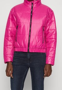 Chaqueta acolchada rosa con cuello alto, que cuenta con una cremallera frontal y bolsillos laterales, fabricada con un material brillante y ligero.