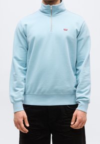 Homme portant un pull à manches longues bleu clair avec une fermeture éclair sur le quart de la longueur, un petit logo rouge sur la poitrine et un pantalon noir.
