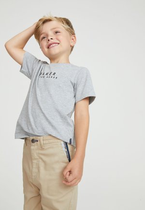 Young blonde boy smiling, wearing gray short-sleeve shirt and beige pants, with one hand behind his head against plain background.