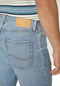 Jeans in denim azzurro chiaro con taglio dritto, dotati di una toppa in pelle beige in vita e cuciture a contrasto. La tasca posteriore presenta un design curvo.