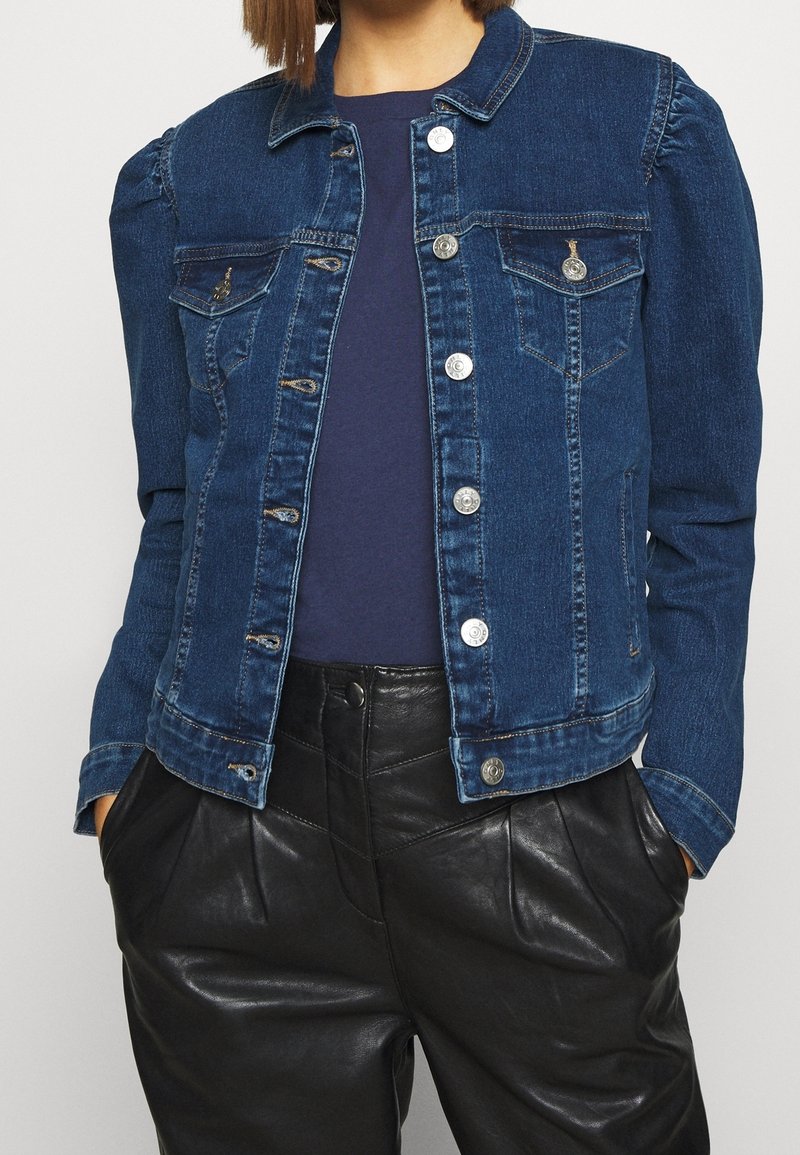 Personne portant une veste en jean bleu foncé avec des boutons argentés et un pantalon en cuir noir taille haute, les mains dans les poches.
