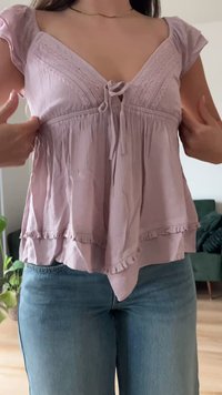 Γυναίκα που ρυθμίζει μια ανοιχτό μωβ μπλούζα με δαντέλα, δεμένη μπροστά, με κυματιστό τελείωμα, φοράει μπλε τζιν σε ένα σαλόνι.