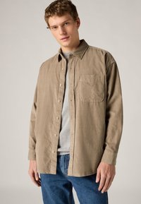 Μπεζ πουκάμισο με μακριά μανίκια και κουμπιά, κατασκευασμένο από μαλακό ύφασμα, με τσέπη στο στήθος και ελαφρώς υφασμάτινη υφή, φορεμένο πάνω από γκρι μπλουζάκι.