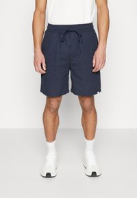 Pantalones cortos azul marino con cintura elástica y cordón. Hechos de tela suave, con bolsillos laterales y un diseño sencillo. Acompañados de zapatillas blancas.