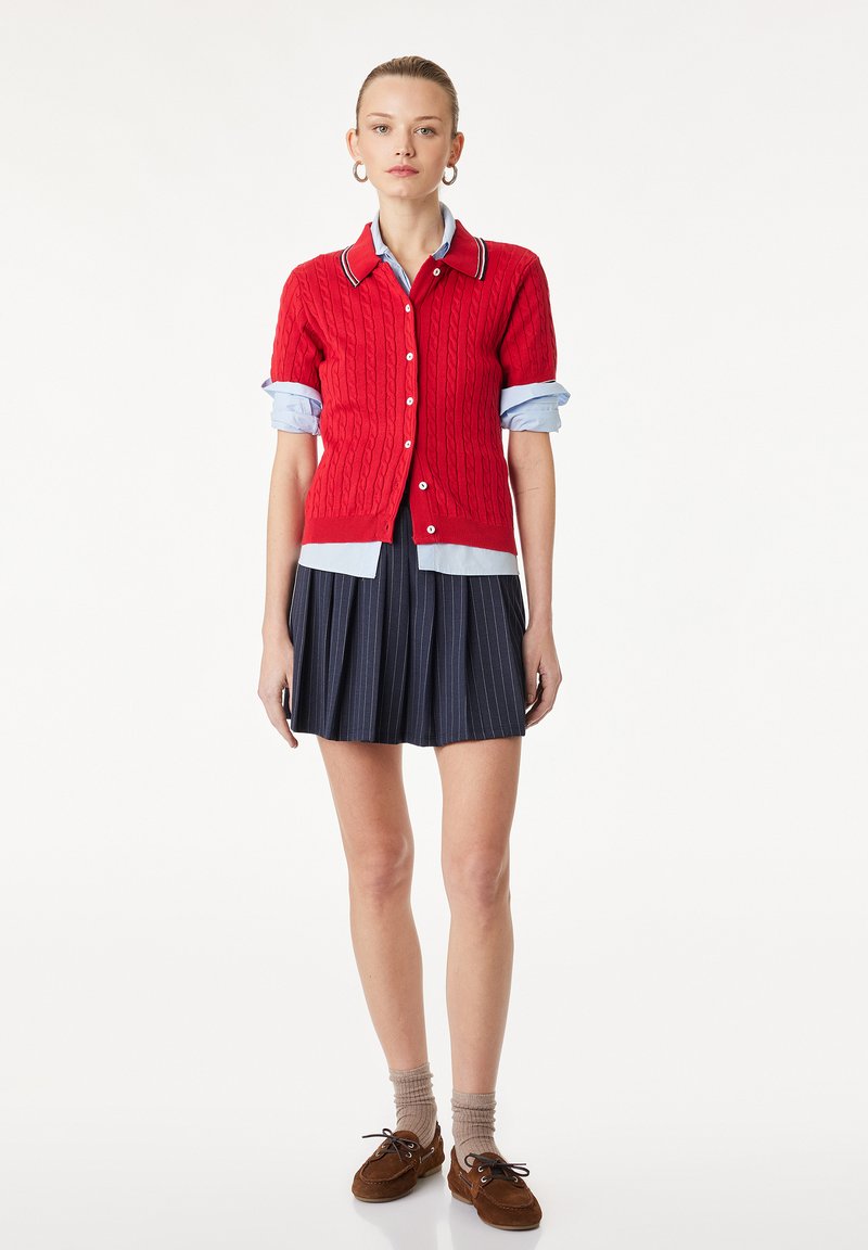 Femme debout portant un cardigan rouge en maille torsadée, une chemise bleue aux manches retroussées, une jupe plissée bleu marine à fines rayures, des chaussures marron et des chaussettes beiges.