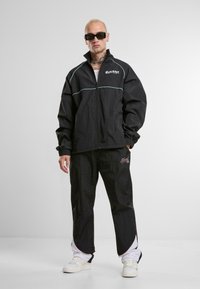Veste coupe-vent noire avec zip, lignes contrastantes bleu clair et pantalon noir assorti présentant un petit logo rose. Porté avec des baskets blanches.