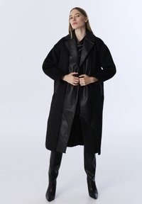 Schwarzer Wollmischungs-Mantel mit Lederakzenten, Knopfverschluss und großem Kragen. Getragen über einem passenden schwarzen Ensemble und kniehohen Stiefeln.