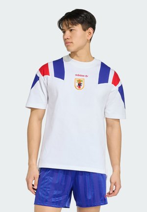Uomo che indossa una maglietta sportiva bianca Adidas con dettagli blu e rossi e pantaloncini sportivi blu, guarda di lato su uno sfondo chiaro uniforme.