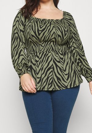 Kvinde iført en grøn og sort zebra-print langærmet bluse med firkantet halsudskæring og blå jeans, vist fra skuldre til hofter.
