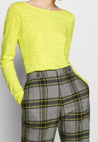 Top à manches longues jaune fluo avec un col bateau, associé à un pantalon à carreaux gris avec des accents verts et une coupe ajustée.