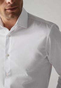 Weißes Hemd mit Knopfleiste aus glattem Stoff und klassischem Kragen, das eine subtile Textur aufweist und zwei sichtbare Knöpfe an der Knopfleiste hat.