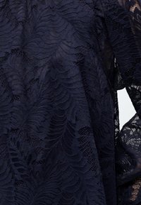 Σκούρο ναυτικό μπλε δαντελένιο ύφασμα με περίπλοκα μοτίβα λουλουδιών και φύλλων, που δείχνει ημιδιάφανη υφή και λεπτή κεντητική.