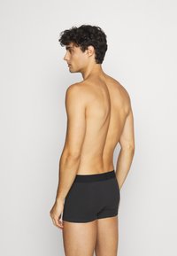 Schwarze Baumwoll-Boxershorts mit elastischem Bund, von hinten betrachtet; das Modell hat einen glatten Rücken und lockiges Haar.