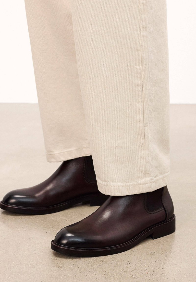 Botine Chelsea din piele maro cu o finisare netedă, vârf rotunjit și panouri laterale elastice, asortate cu pantaloni de culoare crem.