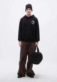 Μαύρο φούτερ με κουκούλα και γραφικό σχέδιο, καφέ παντελόνι με φαρδιά μπατζάκια και στρογγυλή μαύρη τσάντα. Το μοντέλο φοράει μαύρο πλεκτό σκουφάκι με ανάγλυφο κείμενο.