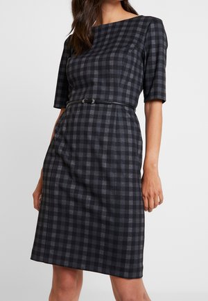 Mujer con vestido a cuadros en negro y gris hasta la rodilla, con mangas hasta el codo y un delgado cinturón negro en la cintura.