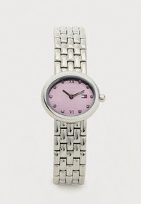 Sudraba metāla saites pulkstenis ar ovālu rozā ciparnīcu, kristāla stundu atzīmēm, romiešu cipariem 12 un 6 un Tommy Hilfiger logo pie 3 stundām.