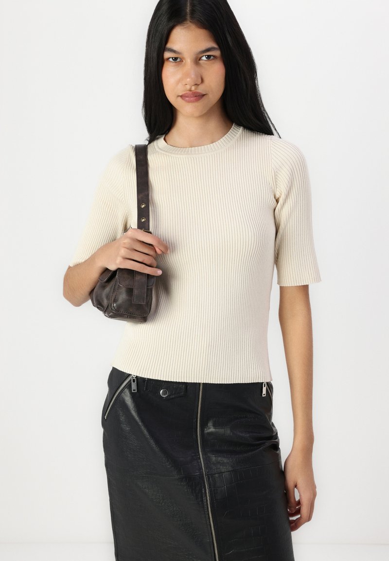 Cremfarbener, gerippter Kurzarm-Top kombiniert mit einem schwarzen Bleistiftrock aus strukturiertem Leder. Das Model hält eine kleine dunkelbraune Handtasche.
