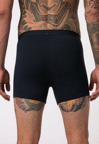 Marineblaue Boxer-Shorts in schmaler Passform, mit einem weichen Taillenbund und Logodetail. Die Haut zeigt lebhafte Tattoo-Muster im Hintergrund.