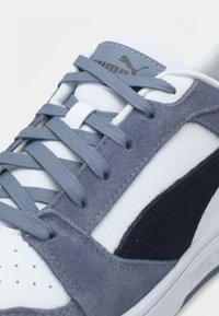 Κοντινή λήψη γκρι και λευκού αθλητικού παπουτσιού Puma με μπλε-γκρι κορδόνια και σουέντ λεπτομέρειες σε λευκό φόντο.