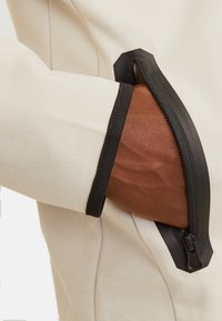 Beige jacksleeve met een zwarte ritszak en een gestructureerde bruine binnenvoering. De hardware van de rits is duidelijk zichtbaar.