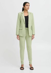 Světle zelený blazer a odpovídající zúžené kalhoty. Blazer má jeden knoflík a ploché kapsy. Pod ním se nachází černý upnutý top, doplněný černými sandály na ploché podrážce.