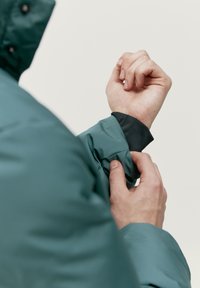 Vodeodolná tyrkysová bunda s hladkým povrchom. Má čierne podšívky na manžetách a nastaviteľné otvory na rukávoch.