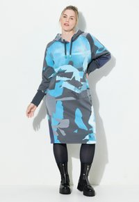Huppari mekko sinisessä ja harmaassa abstraktissa kuosissa, sisältää etutaskun, pitkät hihat ja on tyylitetty mustilla paksupohjaisilla saappailla.