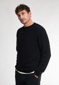 Maglione di lana nero a maglia con scollatura rotonda, maniche lunghe e polsini a costine. Indossato sopra una camicia bianca e abbinato a pantaloni scuri.