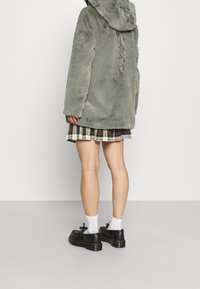 En person iklädd en grön fluffig kappa, rutiga kjolar, vita strumpor och svarta platåskor, som står mot en enkel ljusgrå bakgrund.