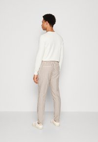 Pantaloni a righe da uomo con strisce verticali beige e blu navy. Presentano un design aderente e sono abbinati a un maglione bianco e a sneakers chiare.
