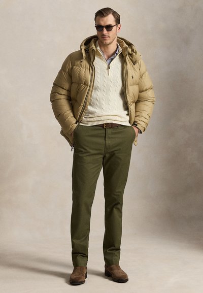 Man wearing tan puffer jacket, cream cable-knit sweater, green pants, brown boots, and sunglasses standing with hands in pockets.