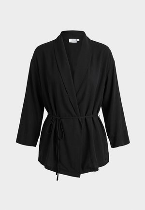 Svart omslagsjakke med trekvartlange ermer, sjalskrage og tynn midjebelte på ensfarget lys grå bakgrunn.