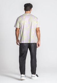 Svetlofialové tričko s žltým botanickým vzorom, krátke rukávy. V páre s tmavými džínsami a bielymi teniskami. Zadný pohľad, jednoduché pozadie.