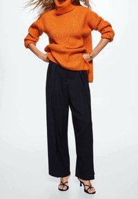 Femme portant un pull à col roulé en maille orange, un pantalon large à rayures noires, et des sandales à talons noires, debout les mains sur les hanches.