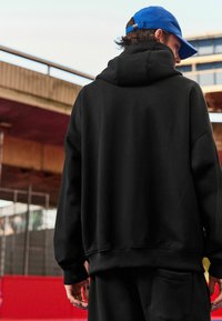Sudadera negra con capucha hecha de un tejido suave, presenta un corte relajado y puños acanalados. Combinada con pantalones de chándal negros a juego.