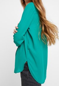 Blusa turchese a maniche lunghe con vestibilità rilassata, caratterizzata da spacco laterale e una texture liscia, abbinata a jeans grigi.